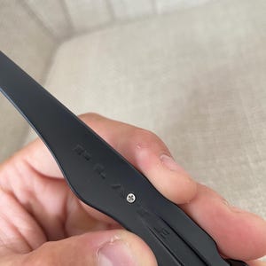 May include: Close-up of a black eyeglass arm with the brand name "POLICE" engraved. The arm is held by a hand and features a small silver screw. The glasses appear to be made of a matte black material.