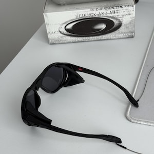 K&ouml;nnte beinhalten: Schwarze Oakley-Sonnenbrille mit dunklen Gl&auml;sern und einem schlanken Design. Die Sonnenbrille liegt auf einer wei&szlig;en Oberfl&auml;che, mit einem silbernen Etui und einer Box mit dem Text "SCIENCE AND ART" im Hintergrund.