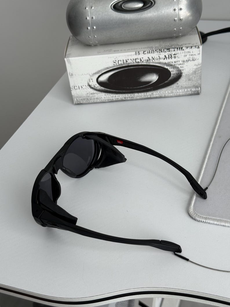 K&ouml;nnte beinhalten: Schwarze Oakley-Sonnenbrille mit dunklen Gl&auml;sern und einem schlanken Design. Die Sonnenbrille liegt auf einer wei&szlig;en Oberfl&auml;che, mit einem silbernen Etui und einer Box mit dem Text "SCIENCE AND ART" im Hintergrund.