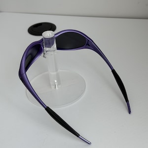 May include: Purple sunglasses with black lenses and black earpieces. The sunglasses are displayed on a clear stand. The frame is a vibrant purple color, and the lenses are dark, providing a sleek, modern look.