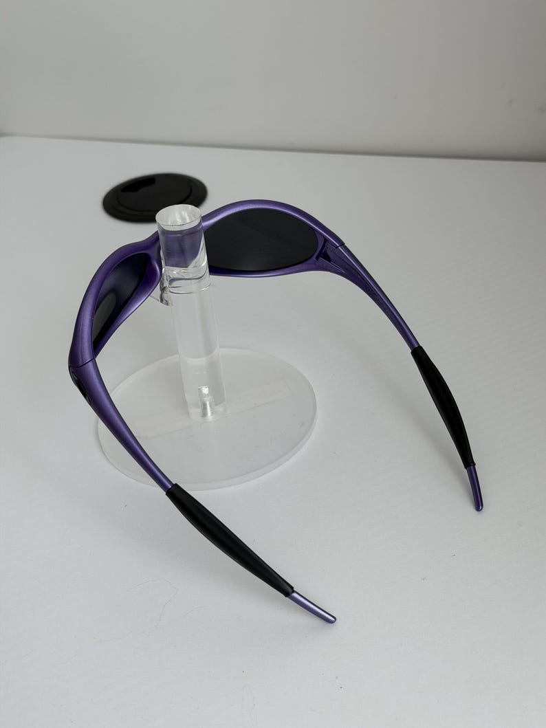 May include: Purple sunglasses with black lenses and black earpieces. The sunglasses are displayed on a clear stand. The frame is a vibrant purple color, and the lenses are dark, providing a sleek, modern look.