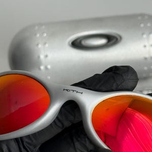 Pu&ograve; includere: Occhiali da sole argentati con lenti rosse vivaci. Gli occhiali hanno un design elegante e moderno con il nome del marchio "KITH" stampato sull'asta. Sullo sfondo &egrave; visibile una custodia argentata.
