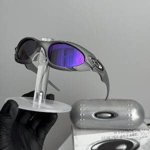 May include: Gray sunglasses with purple lenses are displayed on a clear stand. The sunglasses have a futuristic design, with a silver case and a box with the words "SCIENCE AND ART" visible.