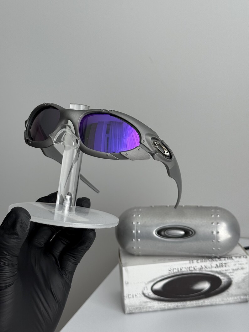 May include: Gray sunglasses with purple lenses are displayed on a clear stand. The sunglasses have a futuristic design, with a silver case and a box with the words "SCIENCE AND ART" visible.