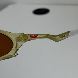 May include: Close-up of sunglasses with a light green frame and brown lenses. The arm of the glasses has a white tip and a red and white "USA Assembled" sticker.