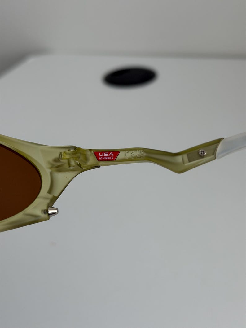 May include: Close-up of sunglasses with a light green frame and brown lenses. The arm of the glasses has a white tip and a red and white "USA Assembled" sticker.