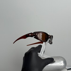 May include: Brown and bronze-colored sunglasses with a sleek, modern design. The sunglasses have a curved frame and tinted lenses. The arms of the sunglasses are a contrasting reddish-brown color. The sunglasses are displayed on a clear stand.