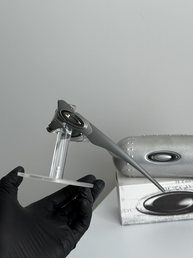 May include: Gray sunglasses with a sleek, modern design. The sunglasses are displayed on a clear stand, with a matching case and box in the background. The Oakley logo is visible on the sunglasses and case.