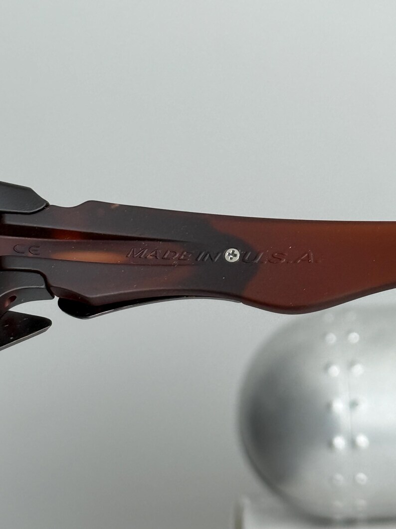 May include: Close-up of the arm of a pair of sunglasses. The arm is brown with a tortoiseshell pattern and the text "MADE IN U.S.A." is visible. A small silver screw is also visible.