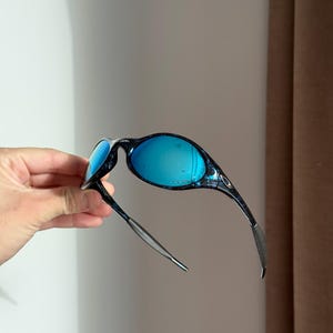 May include: Blue-tinted sunglasses with a black and blue patterned frame. The lenses are a vibrant blue, and the arms are thin and sleek. The sunglasses are held up, showcasing their design.