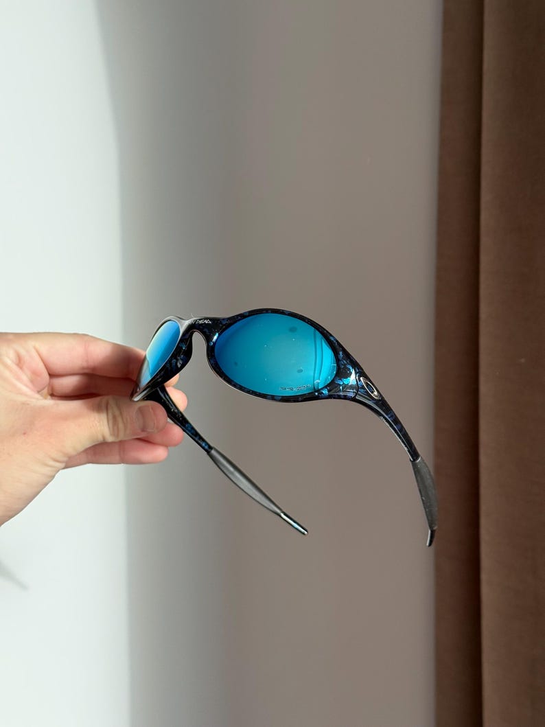 May include: Blue-tinted sunglasses with a black and blue patterned frame. The lenses are a vibrant blue, and the arms are thin and sleek. The sunglasses are held up, showcasing their design.