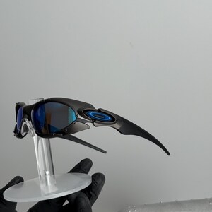 May include: Black sunglasses with blue mirrored lenses are displayed on a clear stand. The sunglasses have a sleek, modern design with curved temples and a circular accent on the frame. The lenses reflect a blue hue.
