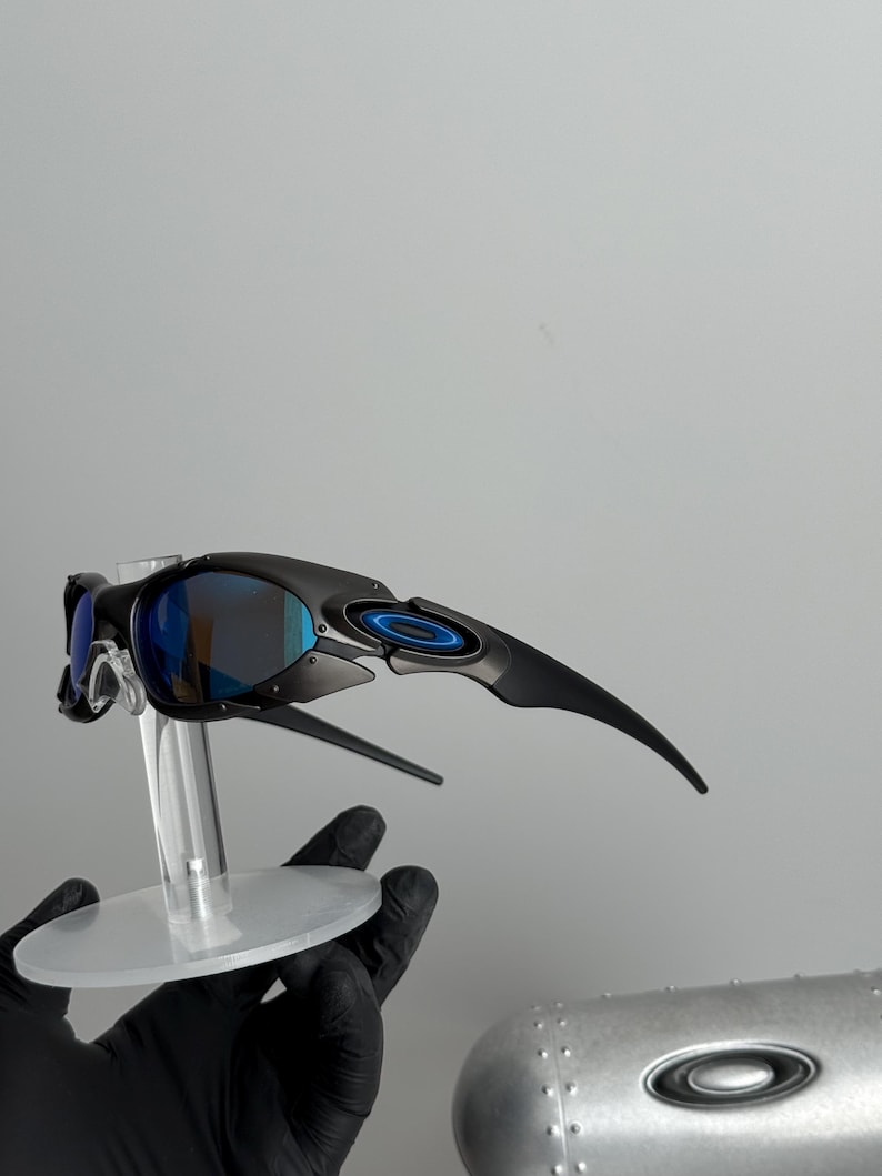 May include: Black sunglasses with blue mirrored lenses are displayed on a clear stand. The sunglasses have a sleek, modern design with curved temples and a circular accent on the frame. The lenses reflect a blue hue.