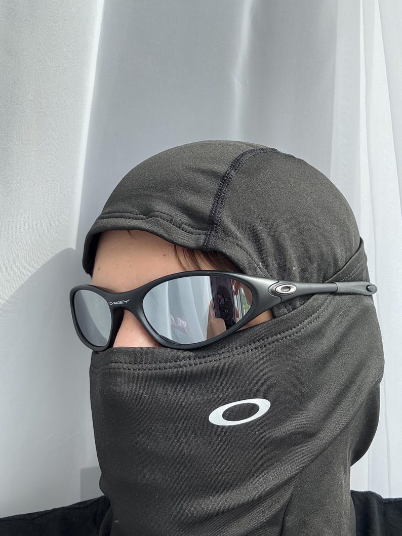 May include: Black sunglasses with mirrored lenses are worn over a dark grey balaclava. The balaclava has a white logo on the front. The sunglasses have a sleek, sporty design.