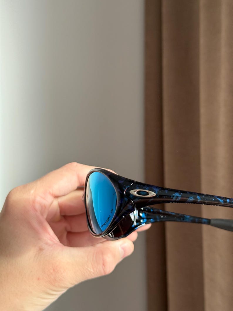 May include: Sunglasses with blue lenses and a blue and black speckled frame. The glasses have a modern design with a dark frame and arms. The lenses reflect a bright blue colour, and the frame has a speckled pattern.