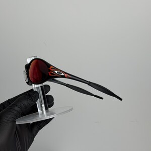 May include: Black and red sunglasses with a flame design on the side. The sunglasses have a dark red lens and are held up by a clear stand. The arms of the sunglasses are black.