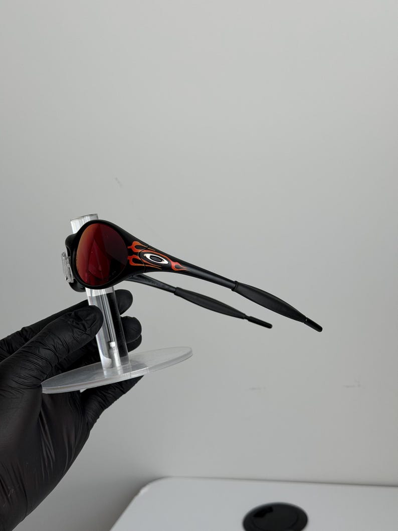 May include: Black and red sunglasses with a flame design on the side. The sunglasses have a dark red lens and are held up by a clear stand. The arms of the sunglasses are black.