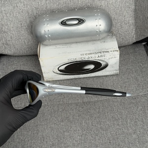 May include: Silver and black sunglasses with a matching silver case and a white box. The sunglasses have a sleek, modern design with a silver frame and black arms. The case is metallic and has a rounded shape.