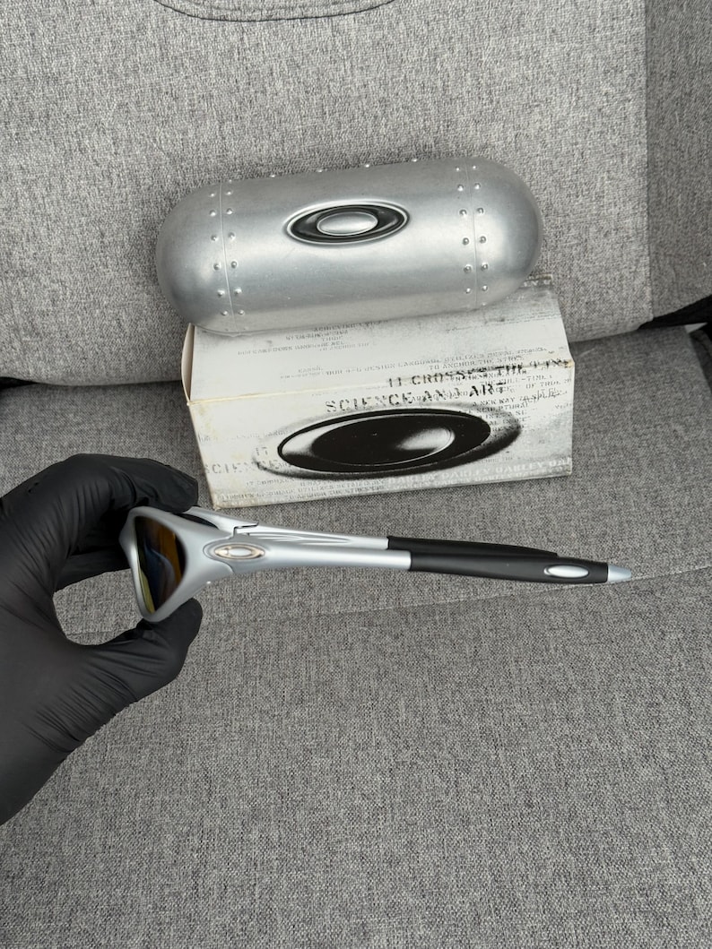 May include: Silver and black sunglasses with a matching silver case and a white box. The sunglasses have a sleek, modern design with a silver frame and black arms. The case is metallic and has a rounded shape.