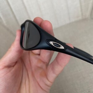 Puede incluir: Gafas de sol negras con lentes oscuras y un acento plateado en la patilla. Las gafas de sol tienen un dise&ntilde;o elegante y moderno y se sostienen en una mano.