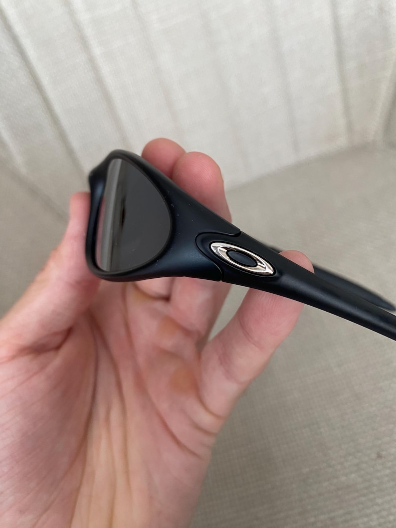 Puede incluir: Gafas de sol negras con lentes oscuras y un acento plateado en la patilla. Las gafas de sol tienen un dise&ntilde;o elegante y moderno y se sostienen en una mano.