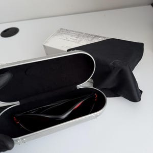 May include: Open silver sunglasses case with black interior, holding black sunglasses with red accents. A black microfiber cloth and a white box are in the background. The sunglasses are designed for sports and outdoor activities.
