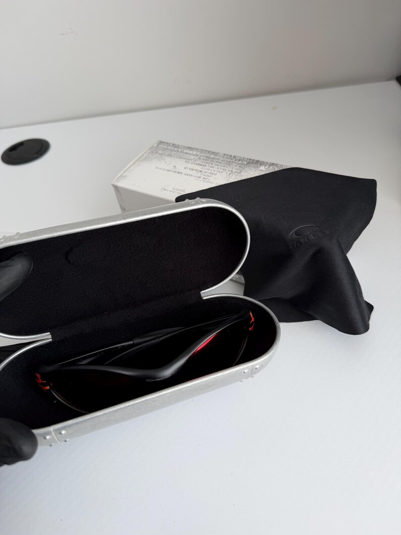 May include: Open silver sunglasses case with black interior, holding black sunglasses with red accents. A black microfiber cloth and a white box are in the background. The sunglasses are designed for sports and outdoor activities.