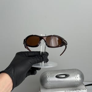 May include: Brown-tinted sunglasses with a dark brown frame, displayed on a clear stand. A silver case and a white box with the text "SCIENCE AND ART" are also visible. The sunglasses have a sporty design.