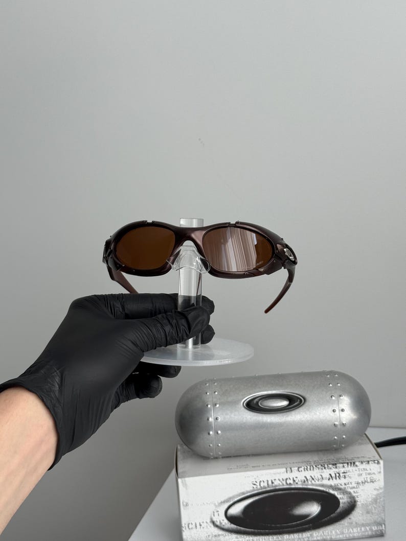 May include: Brown-tinted sunglasses with a dark brown frame, displayed on a clear stand. A silver case and a white box with the text "SCIENCE AND ART" are also visible. The sunglasses have a sporty design.