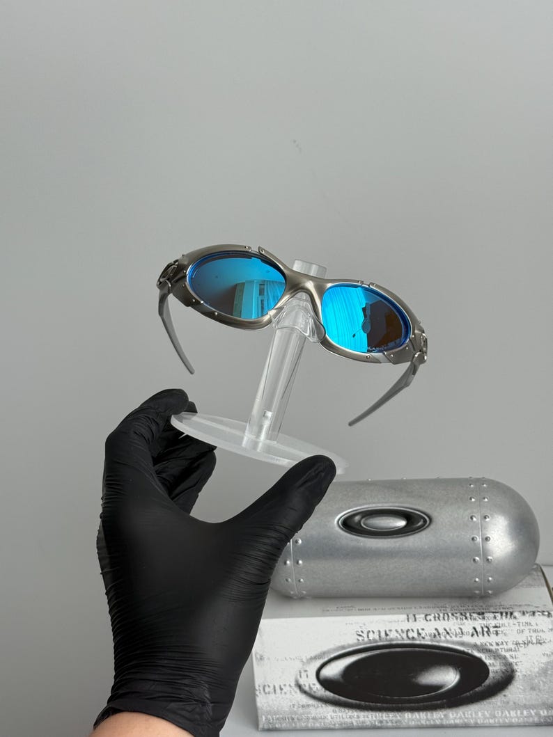 May include: Silver-framed sunglasses with blue mirrored lenses. The sunglasses are displayed on a clear stand, with a silver case and a box with the text "SCIENCE AND ART" visible in the background. The lenses reflect the surrounding environment.