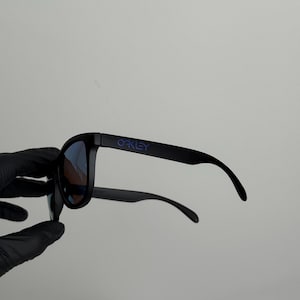 May include: Black sunglasses with blue lenses. The brand name "OAKLEY" is printed in purple on the left arm. The sunglasses are held by a gloved hand against a white background.