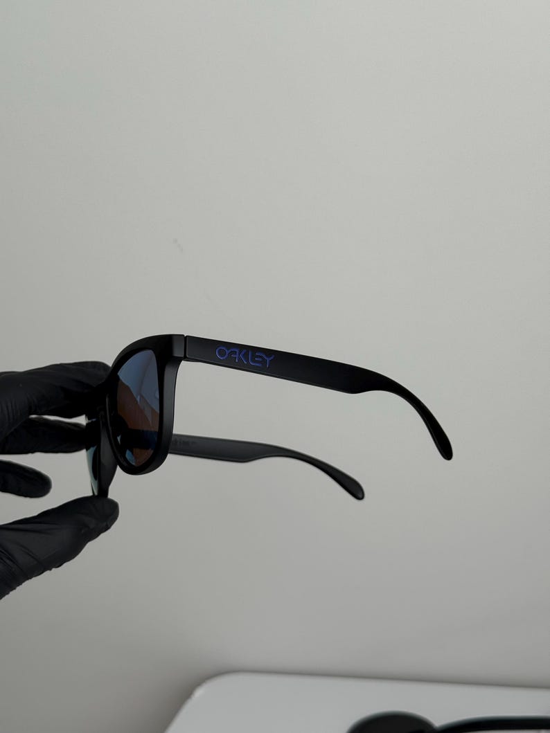 May include: Black sunglasses with blue lenses. The brand name "OAKLEY" is printed in purple on the left arm. The sunglasses are held by a gloved hand against a white background.