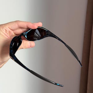 May include: Sunglasses with a dark blue and black marbled pattern on the frames. The lenses are dark, and the temples are a dark grey colour. The sunglasses are held up, showing the design and shape.
