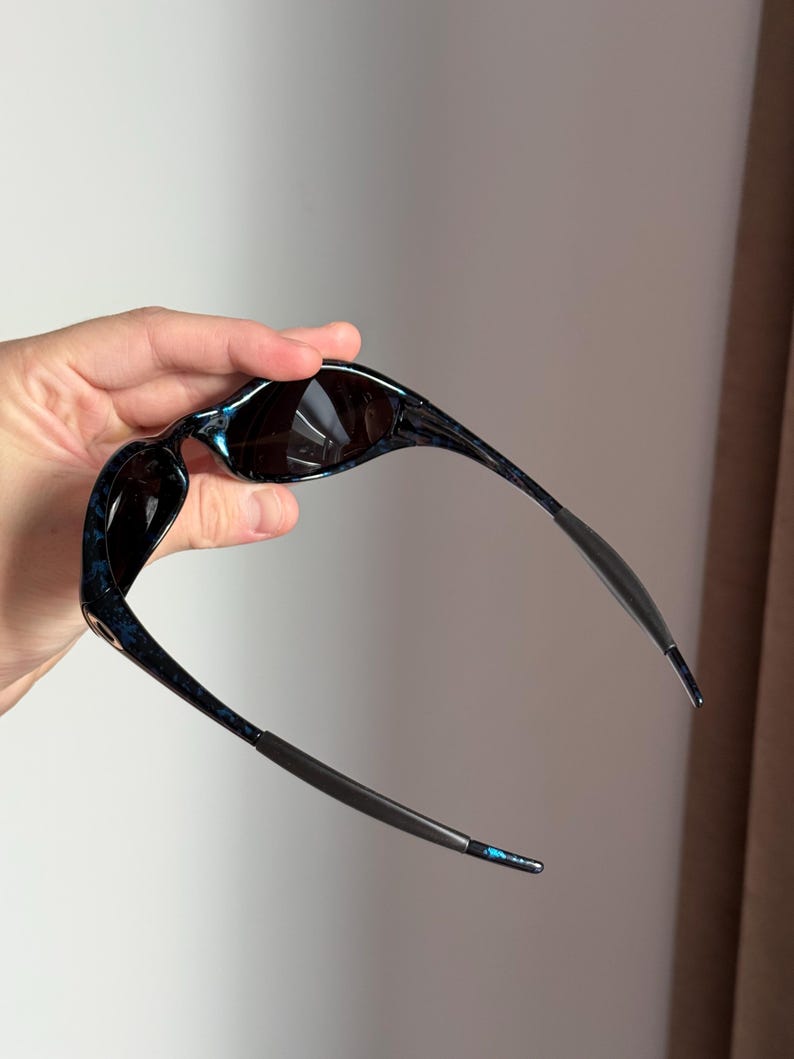 May include: Sunglasses with a dark blue and black marbled pattern on the frames. The lenses are dark, and the temples are a dark grey colour. The sunglasses are held up, showing the design and shape.