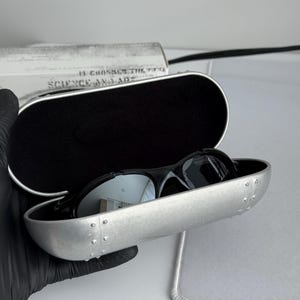 May include: Black sunglasses with reflective lenses rest inside a silver case. The case is open, revealing a black interior. The case has a riveted design and a black border. The text "SCIENCE AND ART" is visible on a white background.