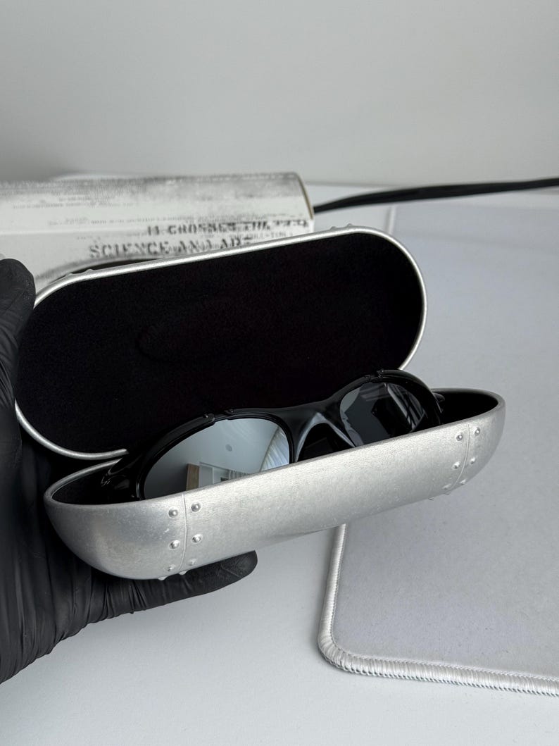 May include: Black sunglasses with reflective lenses rest inside a silver case. The case is open, revealing a black interior. The case has a riveted design and a black border. The text "SCIENCE AND ART" is visible on a white background.