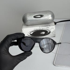 K&ouml;nnte beinhalten: Schwarze Sonnenbrille mit runden Gl&auml;sern und einem schwarzen Rahmen. Die Sonnenbrille wird mit einem silbernen Etui und einer Schachtel mit dem Text "SCIENCE AND ART" pr&auml;sentiert. Die Gl&auml;ser sind dunkel und reflektierend.