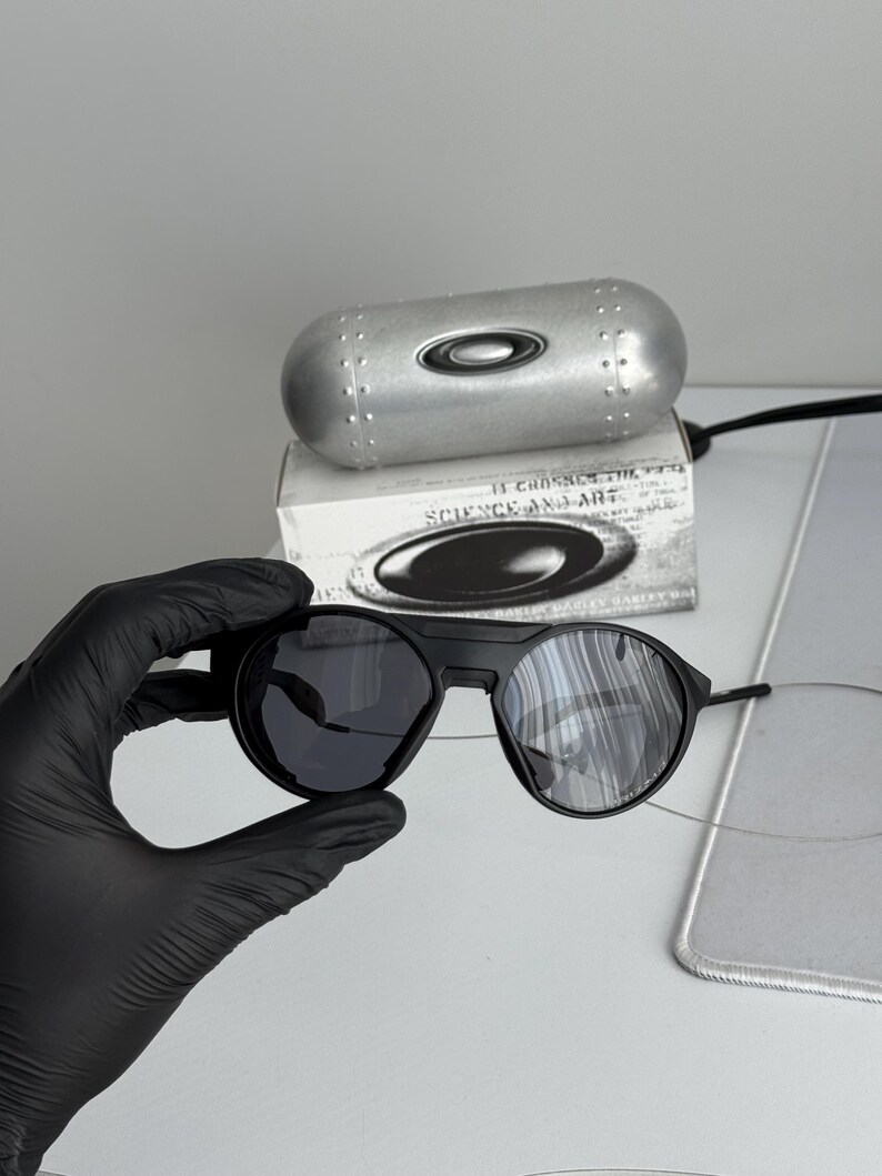 K&ouml;nnte beinhalten: Schwarze Sonnenbrille mit runden Gl&auml;sern und einem schwarzen Rahmen. Die Sonnenbrille wird mit einem silbernen Etui und einer Schachtel mit dem Text "SCIENCE AND ART" pr&auml;sentiert. Die Gl&auml;ser sind dunkel und reflektierend.