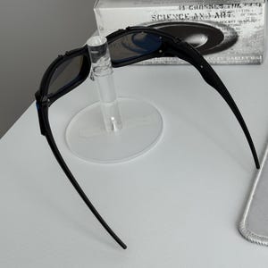 May include: Black sunglasses with blue-tinted lenses are displayed on a clear stand. The sunglasses have a sleek, modern design with a curved frame. The text "SCIENCE AND ART" is visible on the box behind the sunglasses.