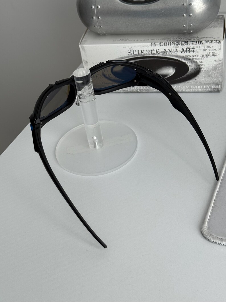 May include: Black sunglasses with blue-tinted lenses are displayed on a clear stand. The sunglasses have a sleek, modern design with a curved frame. The text "SCIENCE AND ART" is visible on the box behind the sunglasses.