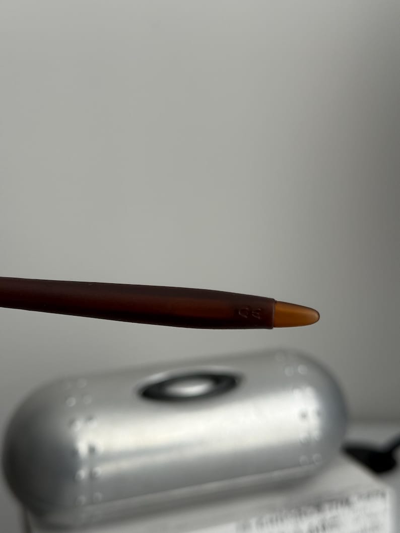 May include: Close-up of a brown and amber-colored eyeglass arm. The arm is slender and tapers to a rounded tip. The Oakley logo is visible on the silver case in the background.