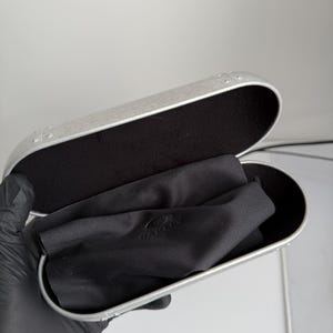 May include: Open silver-colored eyeglasses case with a black interior and a black cleaning cloth inside. The case has a rounded rectangular shape and a metallic finish. The cloth has the brand name "OAKLEY" printed on it.