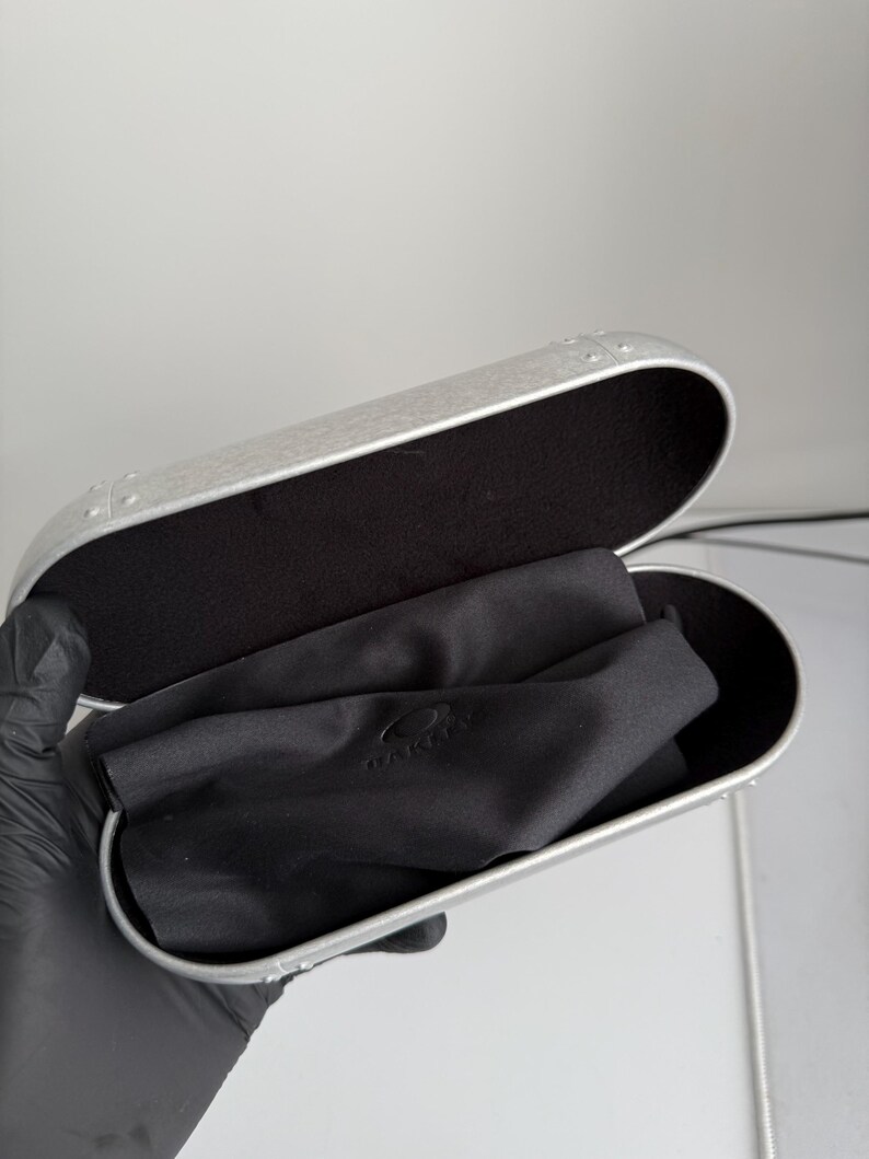 May include: Open silver-colored eyeglasses case with a black interior and a black cleaning cloth inside. The case has a rounded rectangular shape and a metallic finish. The cloth has the brand name "OAKLEY" printed on it.