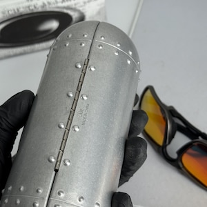 May include: A silver-colored, riveted sunglasses case with the text "CASE MADE IN CHINA" on the hinge. The case is open, revealing a pair of sunglasses with orange-tinted lenses. The case is held by a gloved hand.