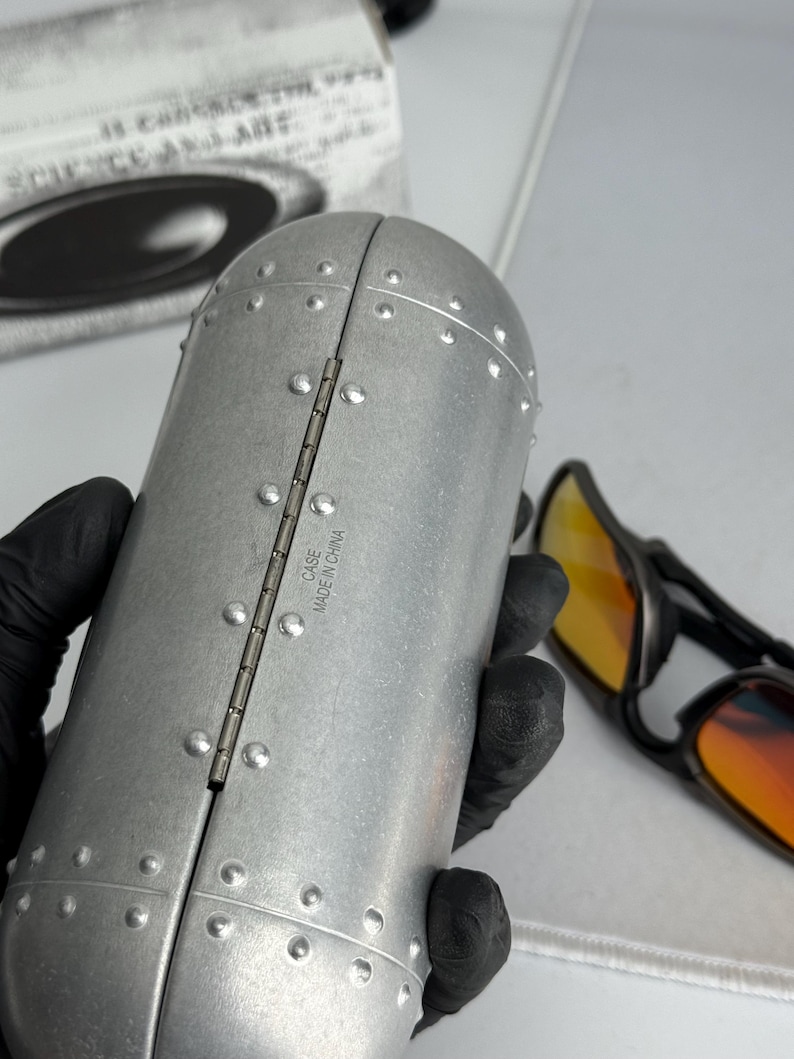May include: A silver-colored, riveted sunglasses case with the text "CASE MADE IN CHINA" on the hinge. The case is open, revealing a pair of sunglasses with orange-tinted lenses. The case is held by a gloved hand.