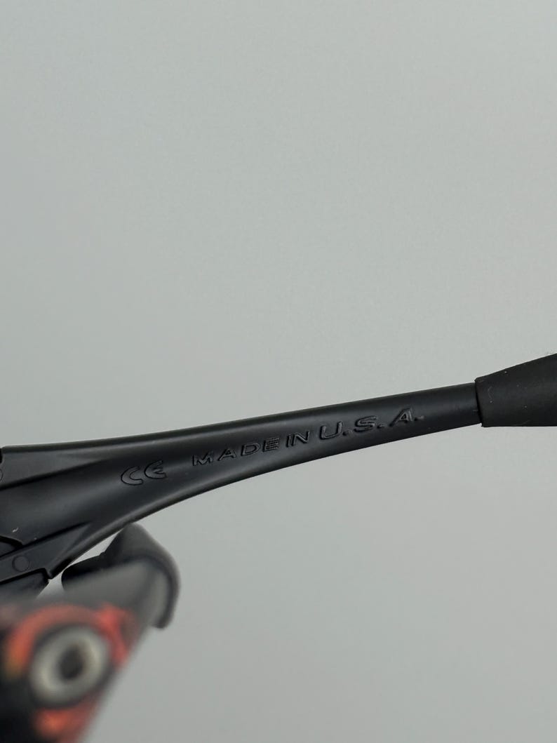 May include: Close-up of black sunglasses with the text "MADE IN U.S.A." printed on the arm. The sunglasses have a sleek, modern design with a matte finish. The frame is black, and the lenses are not visible.