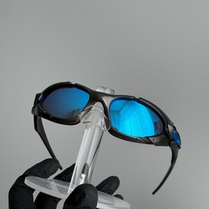 May include: Black sunglasses with blue reflective lenses. The sunglasses are held on a clear display stand. The lenses reflect the surrounding environment. The frames are black and have a sporty design.