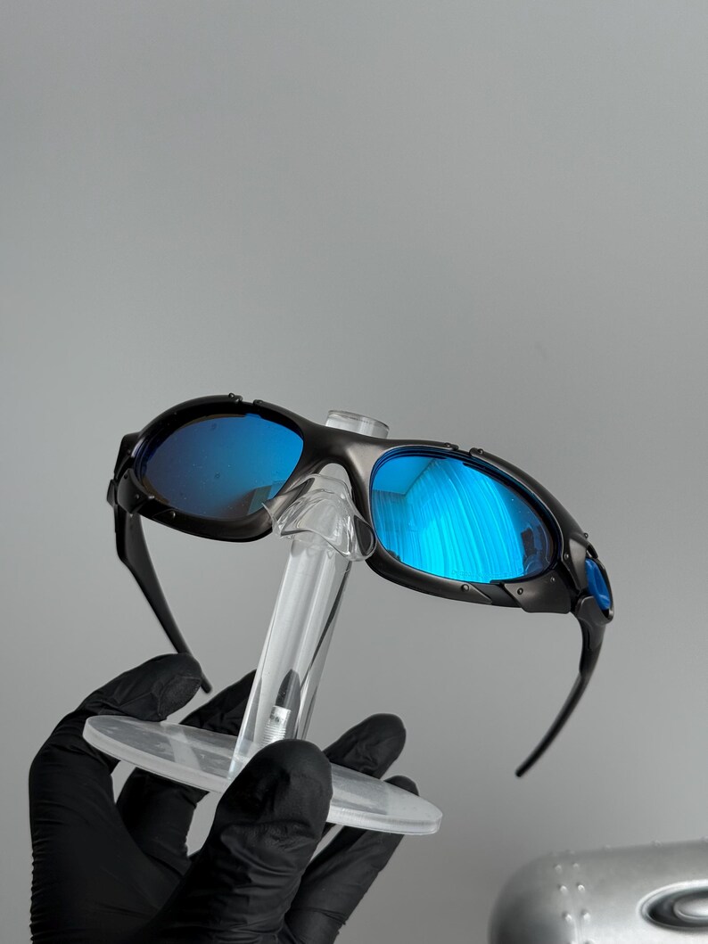 May include: Black sunglasses with blue reflective lenses. The sunglasses are held on a clear display stand. The lenses reflect the surrounding environment. The frames are black and have a sporty design.