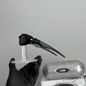 May include: Black sunglasses with a sleek, aerodynamic design, displayed on a clear stand. A silver case and a white box with the text "SCIENCE AND ART" are also visible.