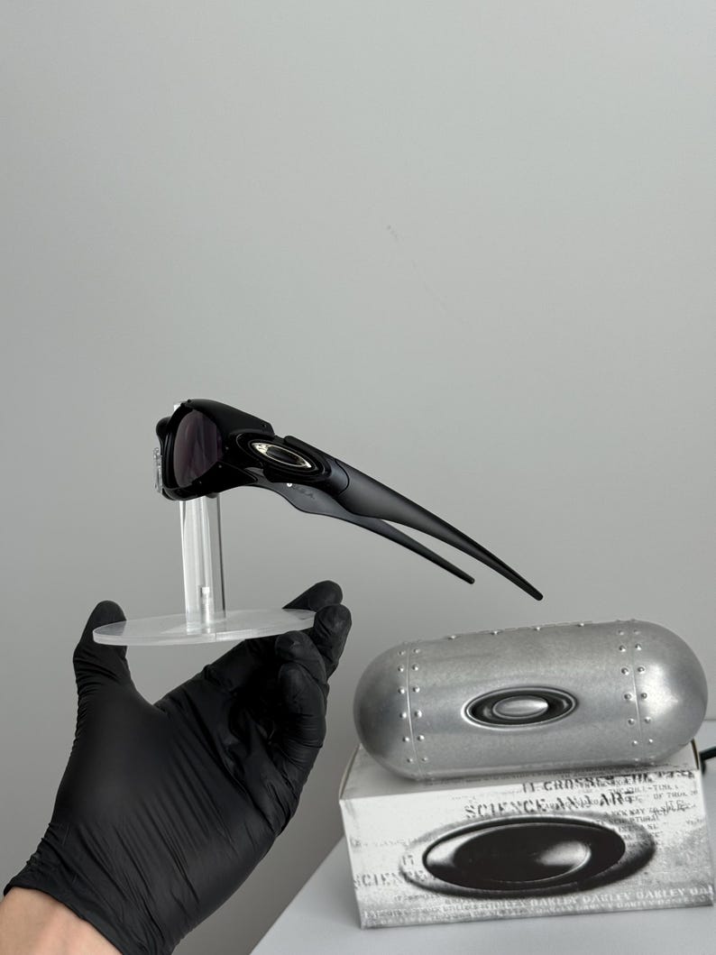 May include: Black sunglasses with a sleek, aerodynamic design, displayed on a clear stand. A silver case and a white box with the text "SCIENCE AND ART" are also visible.